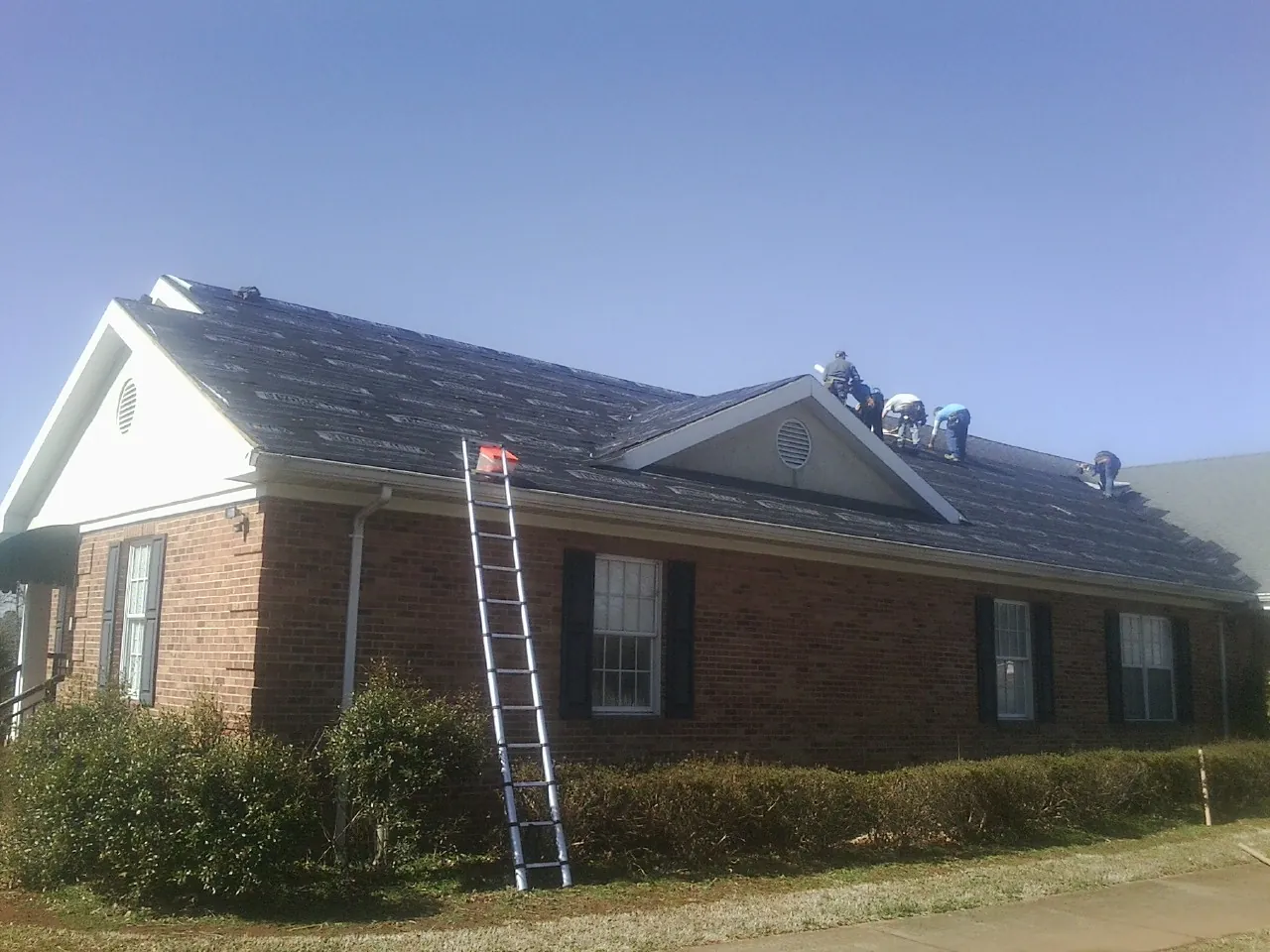 Crew tearing shingles off a brick ranch roof with a ladder set — Emergency Roof Repair work by our crew in Saint Hedwig