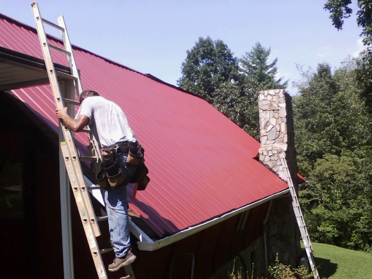 Roofer on a ladder beside a red metal roof and stone chimney — Soffit & Fascia Repair by Roofing Brothers in Saint Hedwig